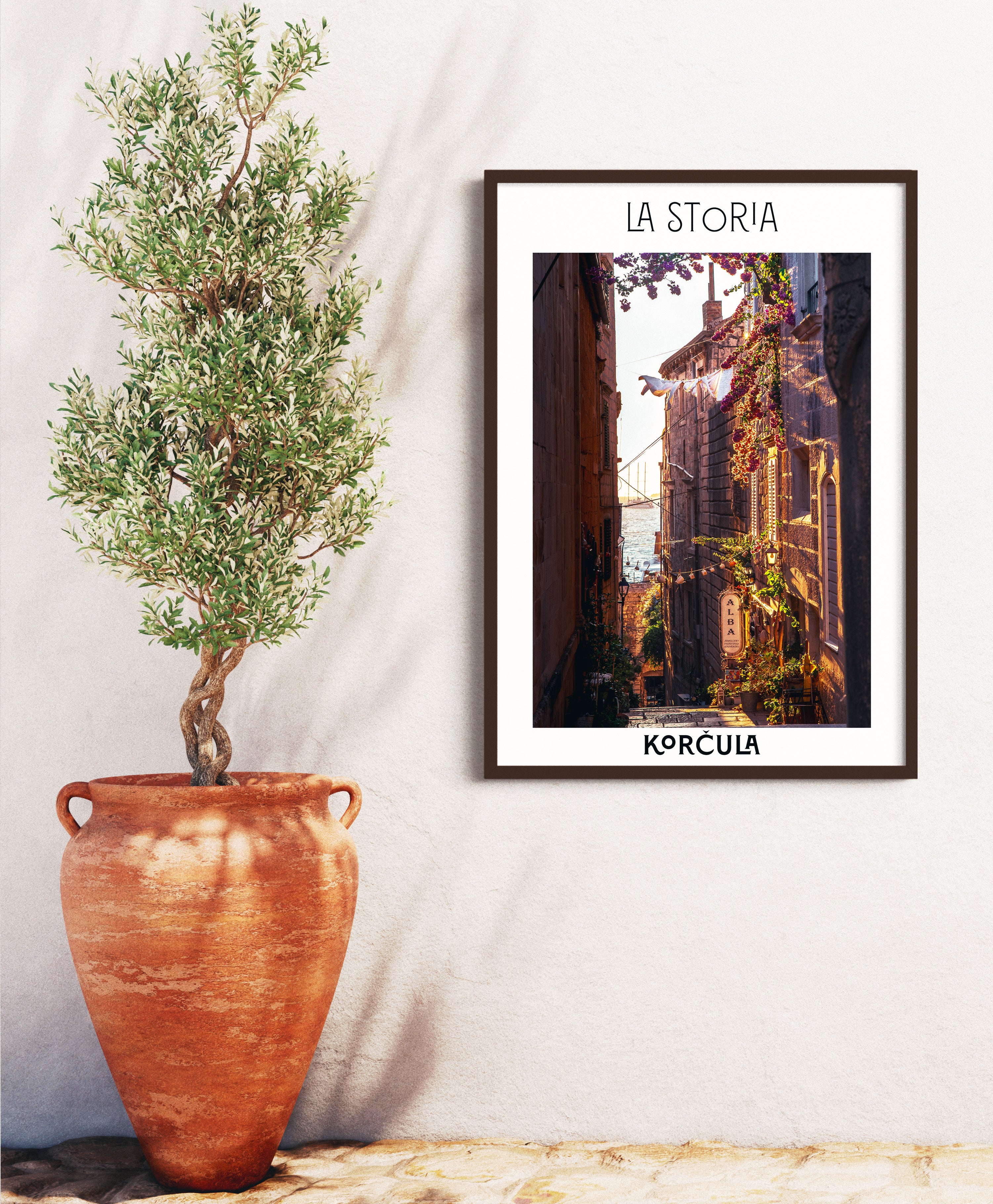 Framed artwork titled 'LA STORIA' by Korcula next to a potted olive tree on a white wall.