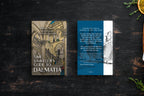 A book titled 'The Time Traveler's Guide to Dalmatia' shown alongside a matching travel card or insert on a dark wooden surface. The book cover features an illustration of a historic interior with arches and stairs, while the insert has a blue vertical band with white text and a black-and-white illustration of a woman.
